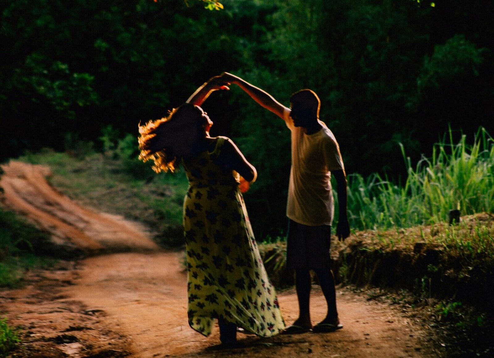 A couple gracefully dances on a sunlit dirt road surrounded by lush greenery in Saquarema, Brazil.